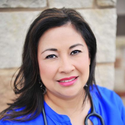 Woman in a blue top with a stethoscope, smiling against a stone wall.