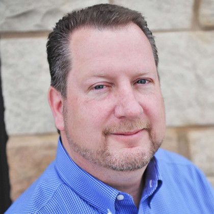 Headshot of a man in a blue shirt smiling slightly against a stone wall background