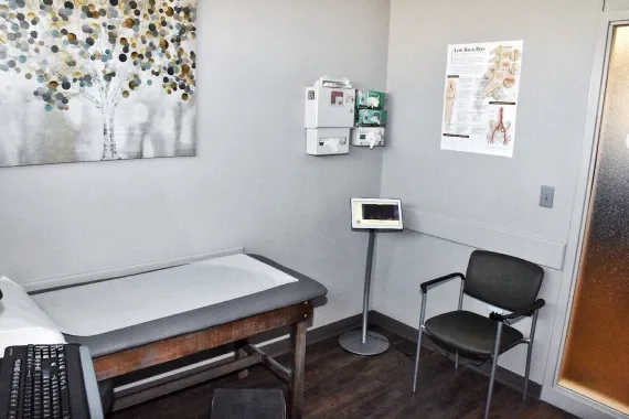 Medical exam room with exam table, chair, monitor, wall posters, and sink cabinet
