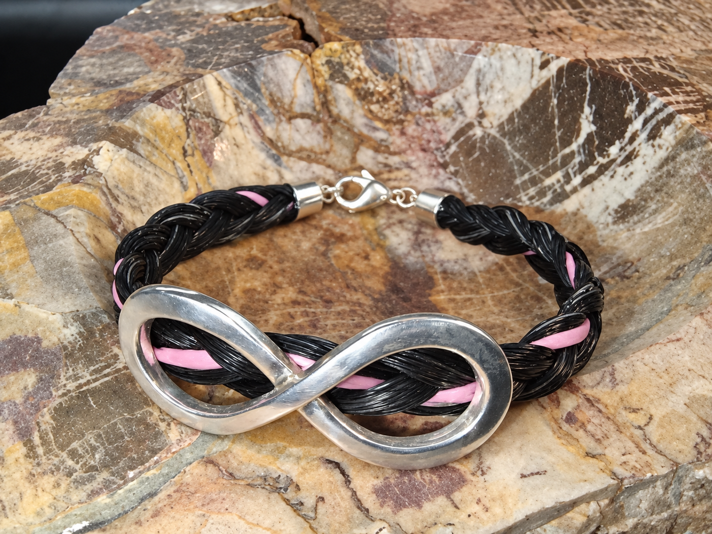 Charm infini argenté sur un bracelet tressé noir avec des touches de rose, posé sur une surface en pierre