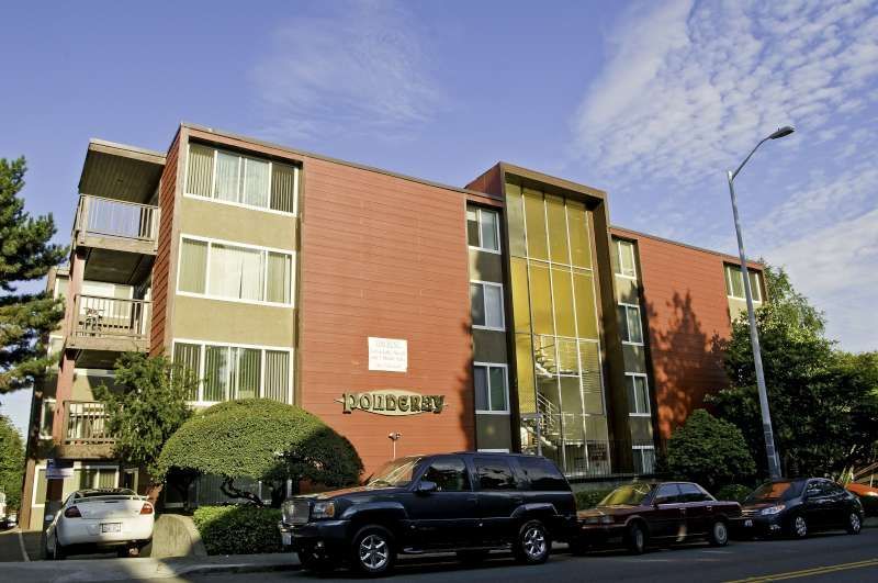 Apartment building with orange brick and green balconies, parked cars along the street under a blue sky.