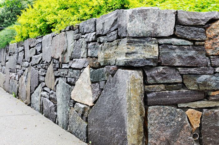 A stone wall along a sidewalk with trees in the background.