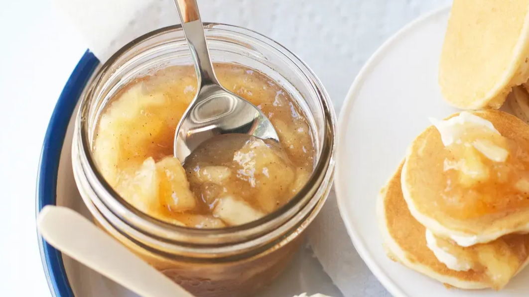 Een potje appelcompote met een lepel en pannenkoeken besprenkeld met siroop.