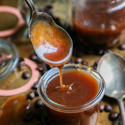 Karamelsaus druipt van een lepel in een pot, omringd door koffiebonen en andere potten.