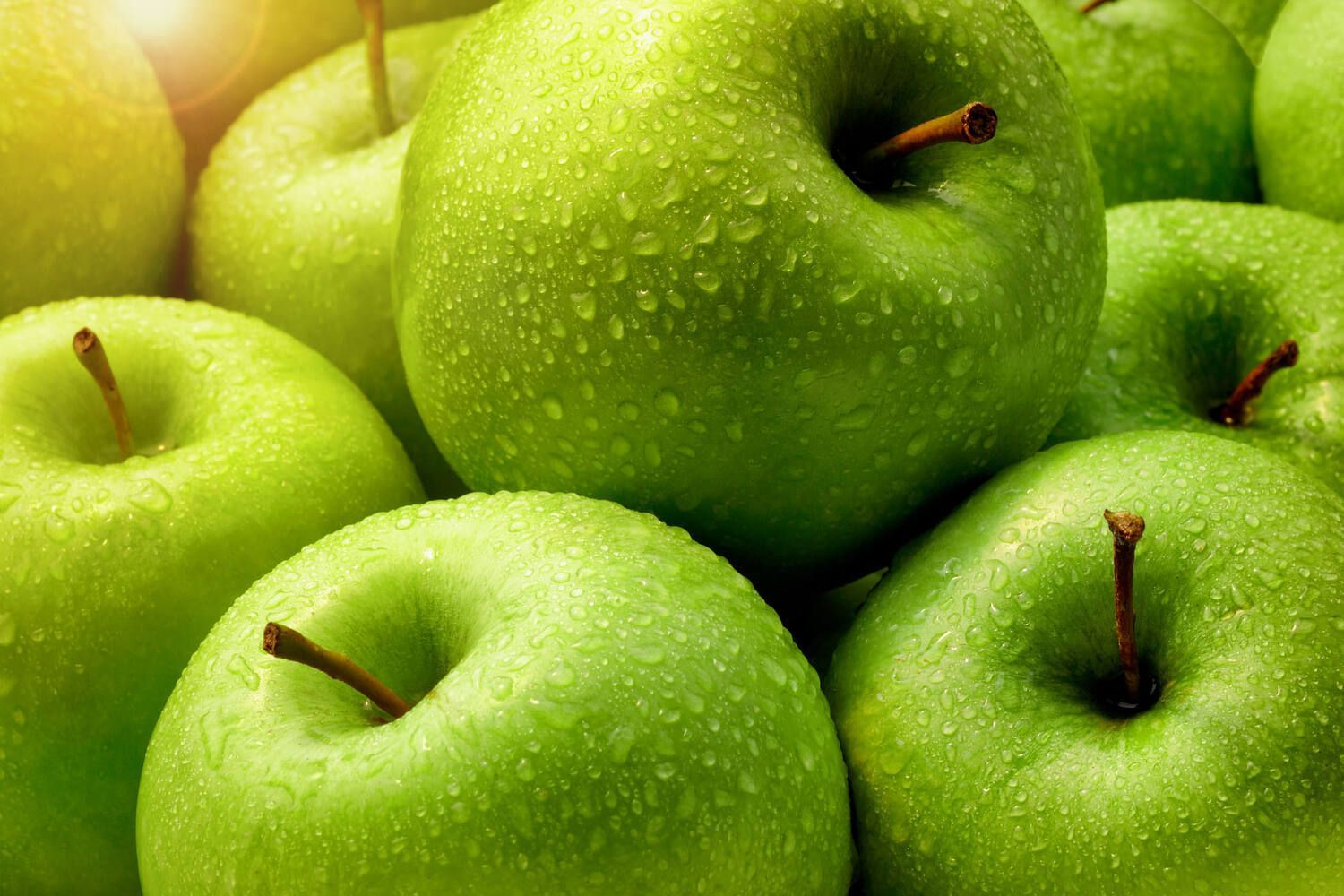 Close-up van een stapel verse, groene appels met waterdruppels, waarbij de steeltjes zichtbaar zijn.
