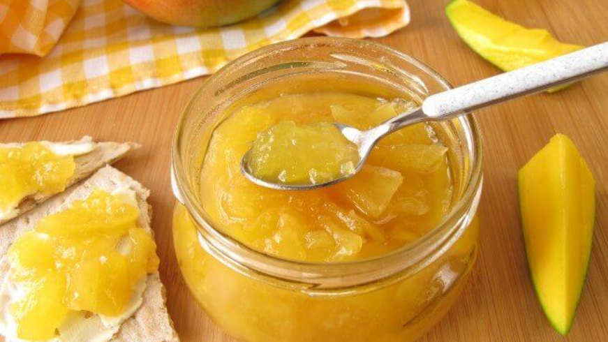 Mangojam in een pot, een lepel die in de jam wordt gedoopt, plakjes mango en brood met jam op een houten ondergrond.