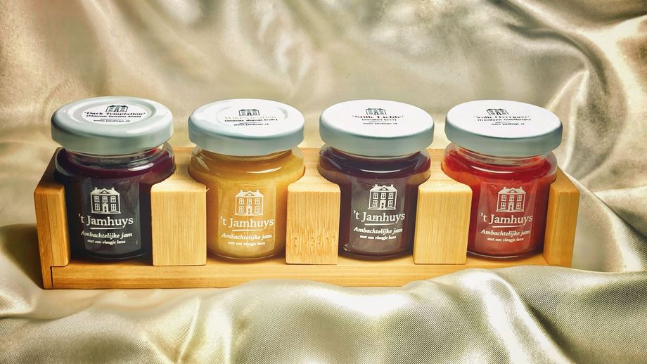 Vier kleine jampotjes in een houten houder op een glanzende gouden doek, met witte deksels en etiketten.