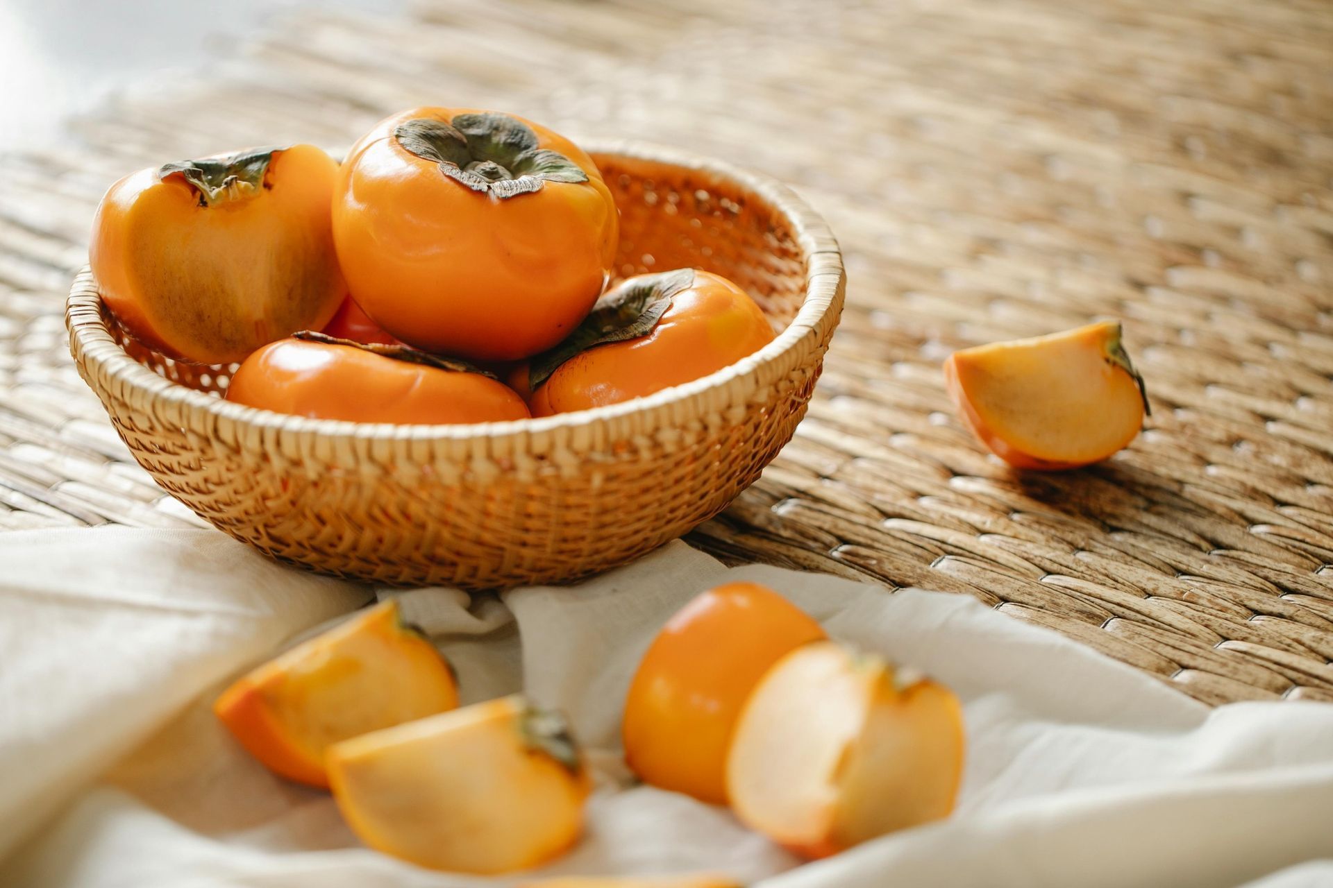 Een mand met rijpe kaki's, sinaasappels en schijfjes fruit op een geweven mat.