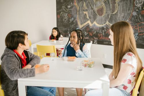 People seated around a white table in a bright room, chatting and eating with a chalkboard wall behind them
