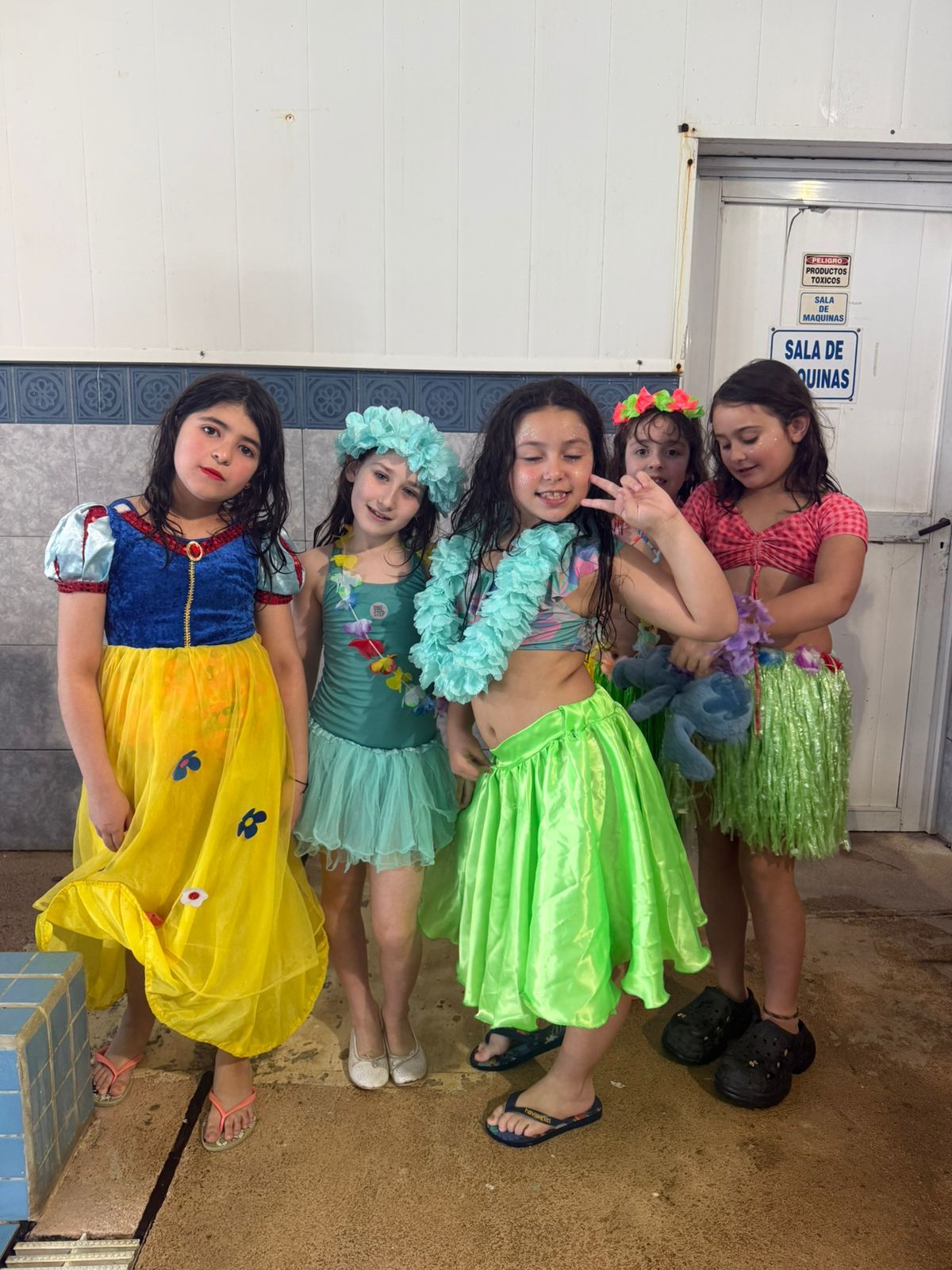 Cinco niños disfrazados posan junto a una piscina. Se ven trajes de Blancanieves, sirena y hula hula. Sonríen.