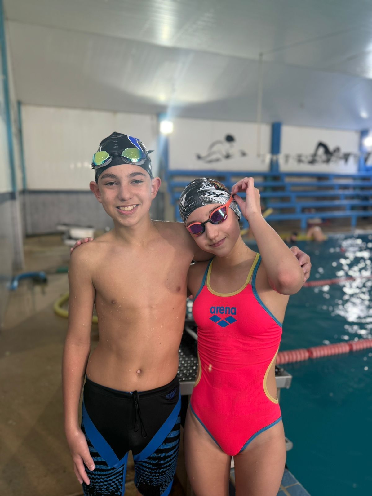Dos nadadores en una piscina, sonriendo y posando. La chica lleva un bañador rosa y gafas protectoras. El chico lleva traje de baño.