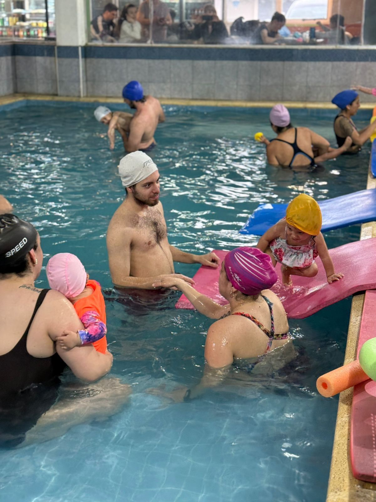 Personas con niños en una piscina, algunos de ellos con gorro de baño y utilizando dispositivos de flotación.