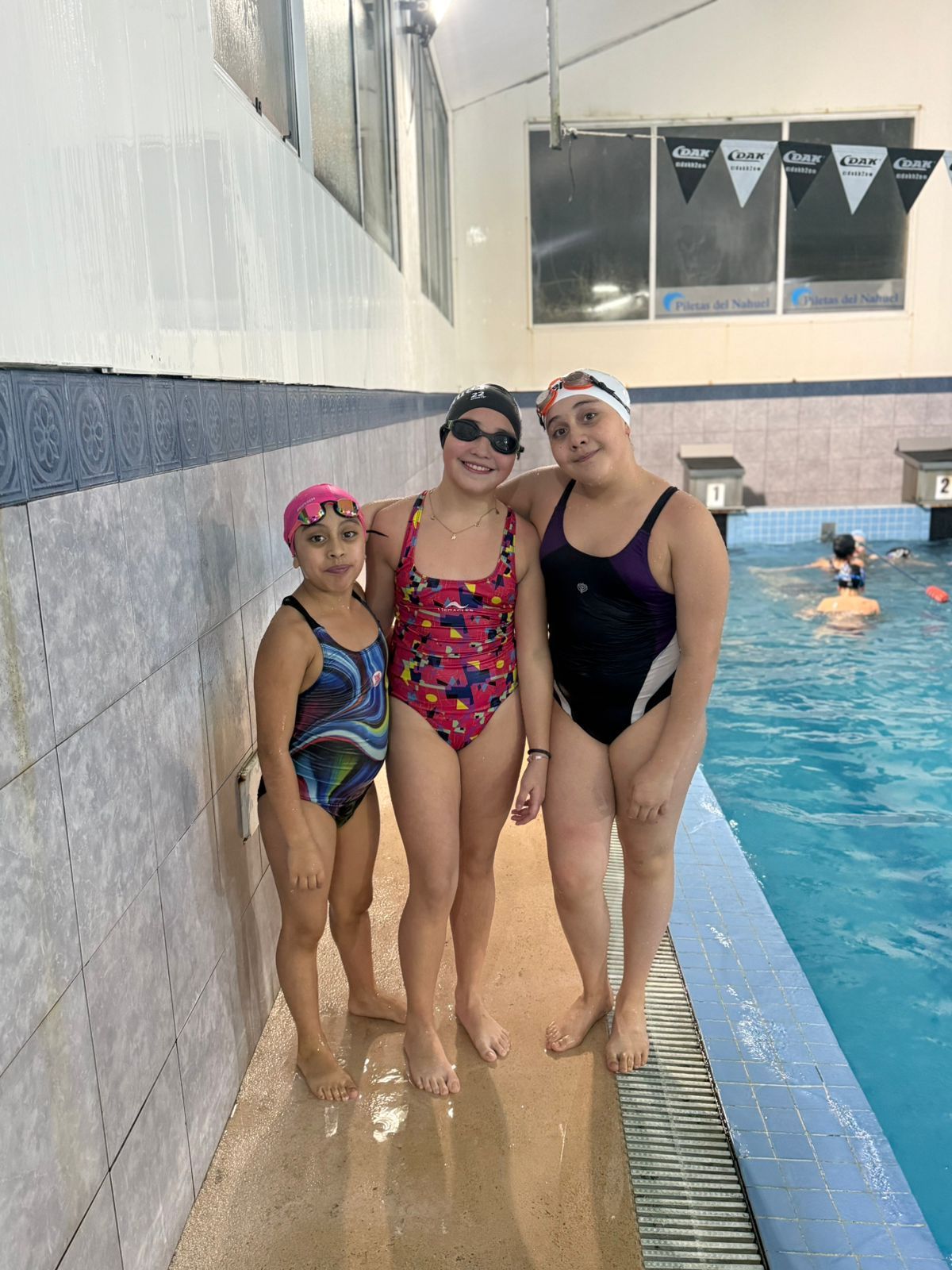 Tres personas en traje de baño posan junto a una piscina. Dos llevan gorro de baño. Una sonríe y abraza a las demás.