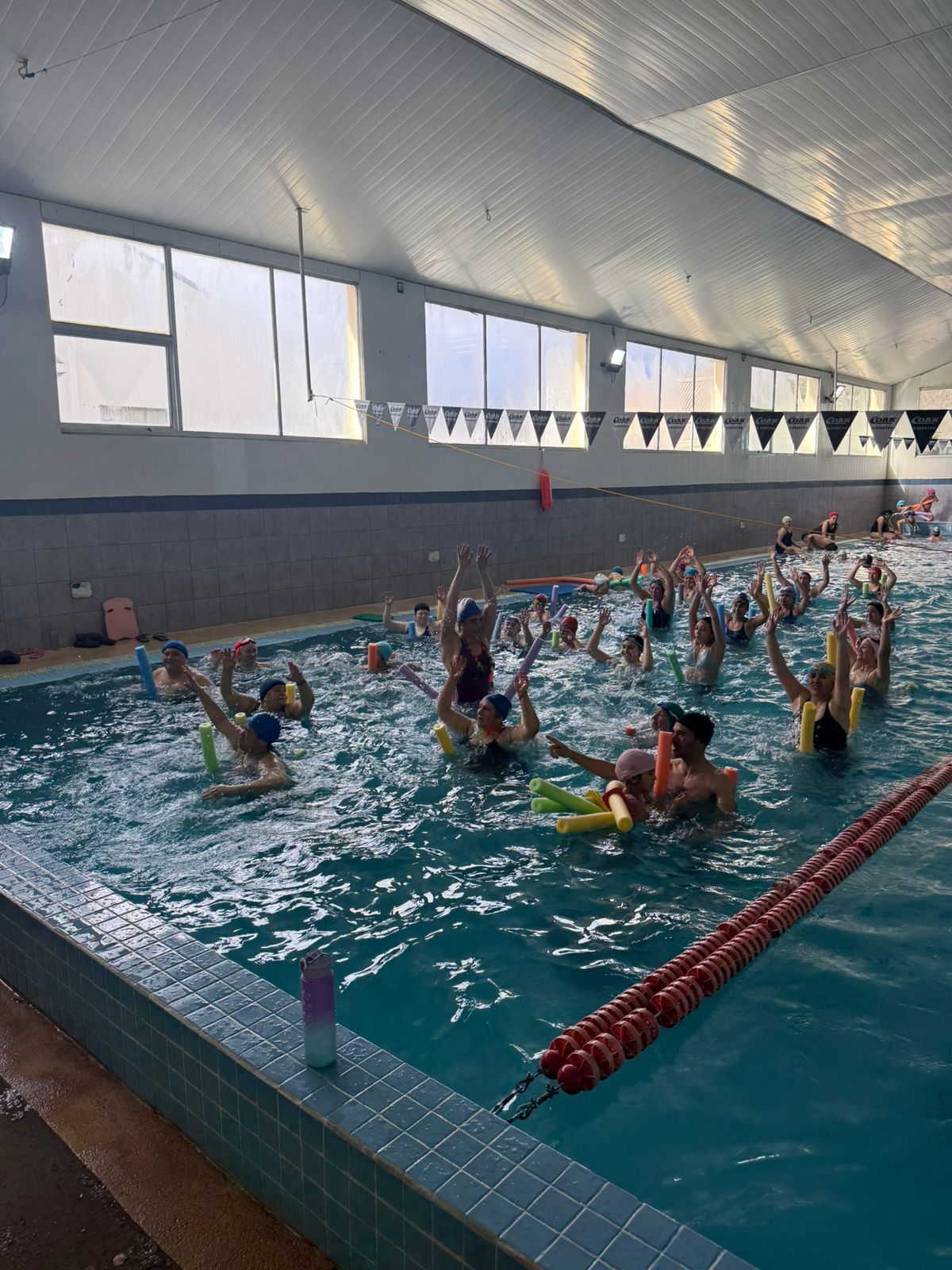Personas en una piscina, algunas con flotadores, levantando las manos. Piscina cubierta.