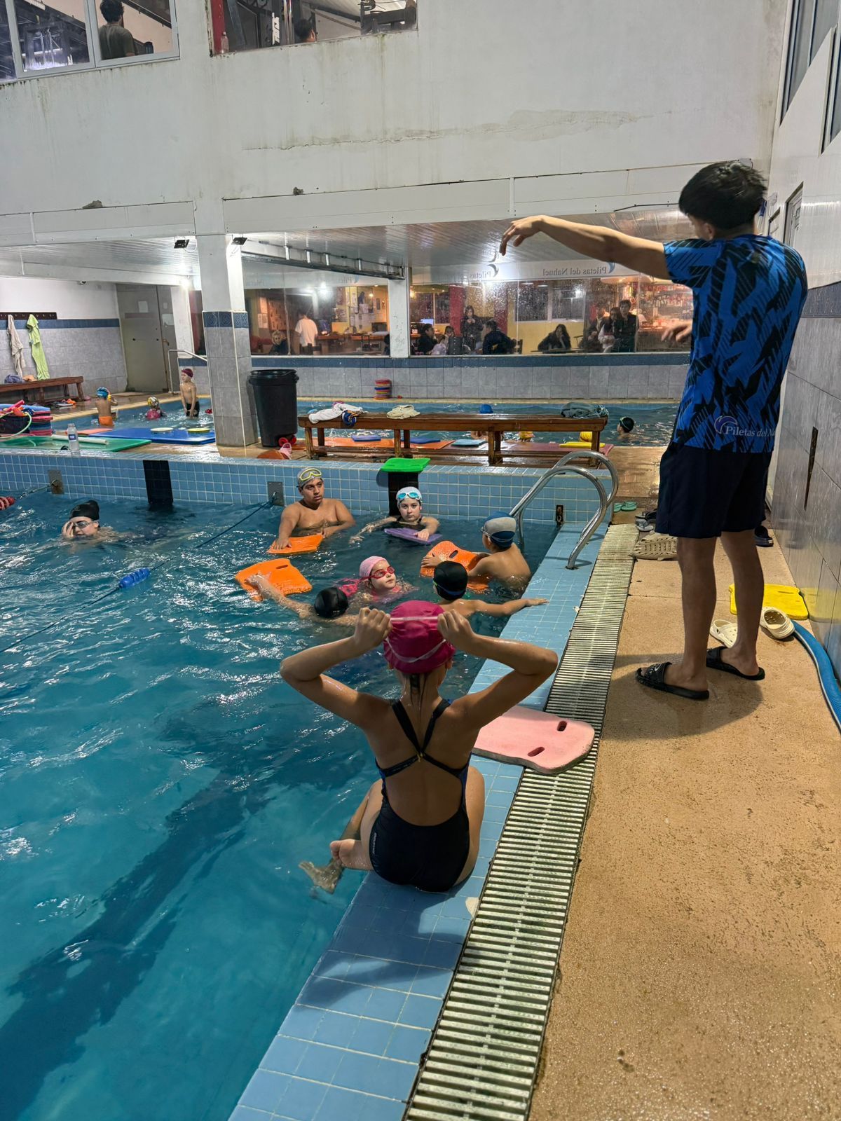 Un instructor de natación señala a los niños mientras aprenden a nadar en una piscina. Una niña en traje de baño se ajusta las gafas.