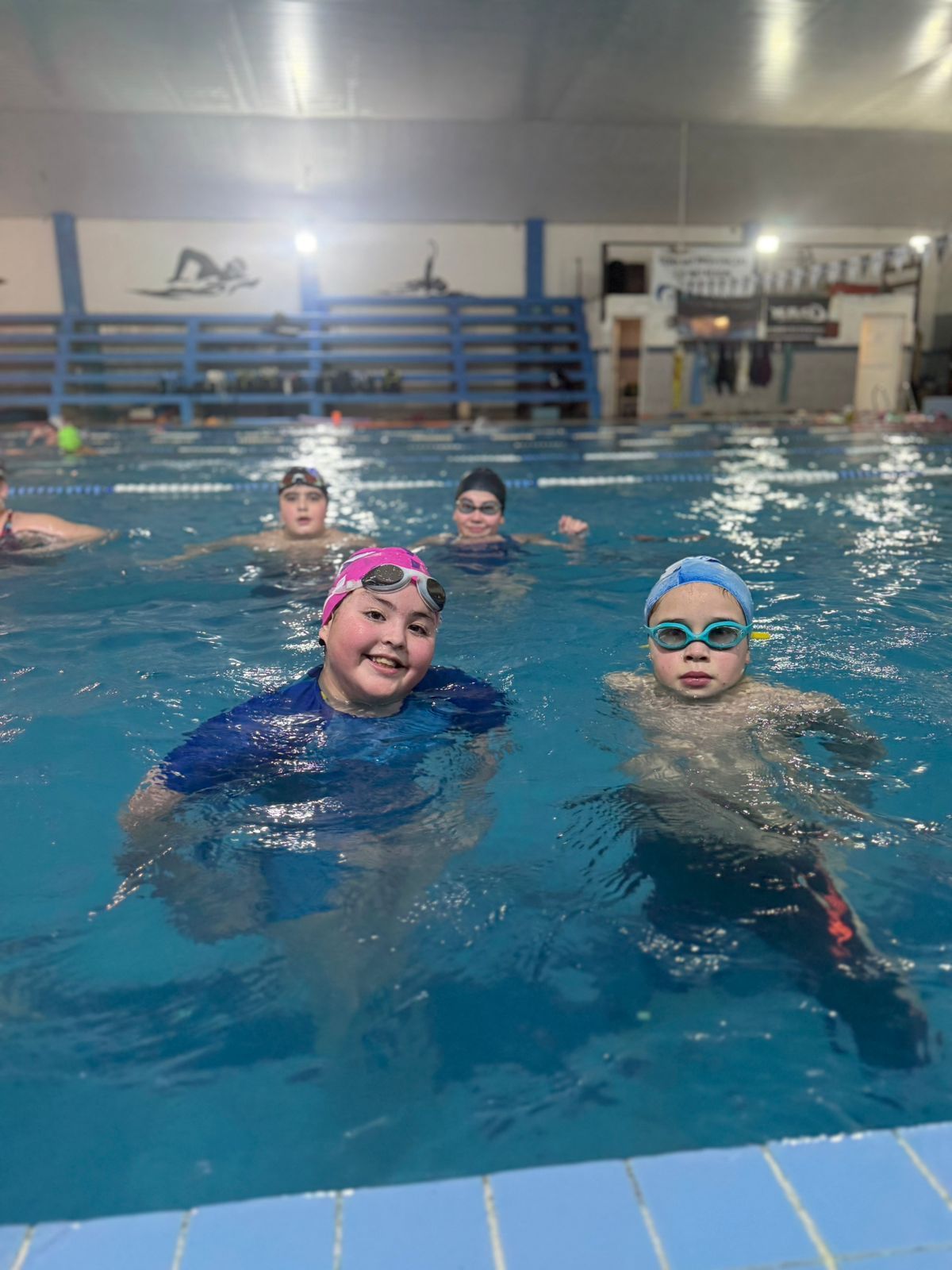 Niños en una piscina, algunos con gorro y gafas de natación, sonriendo a la cámara. Piscina cubierta.