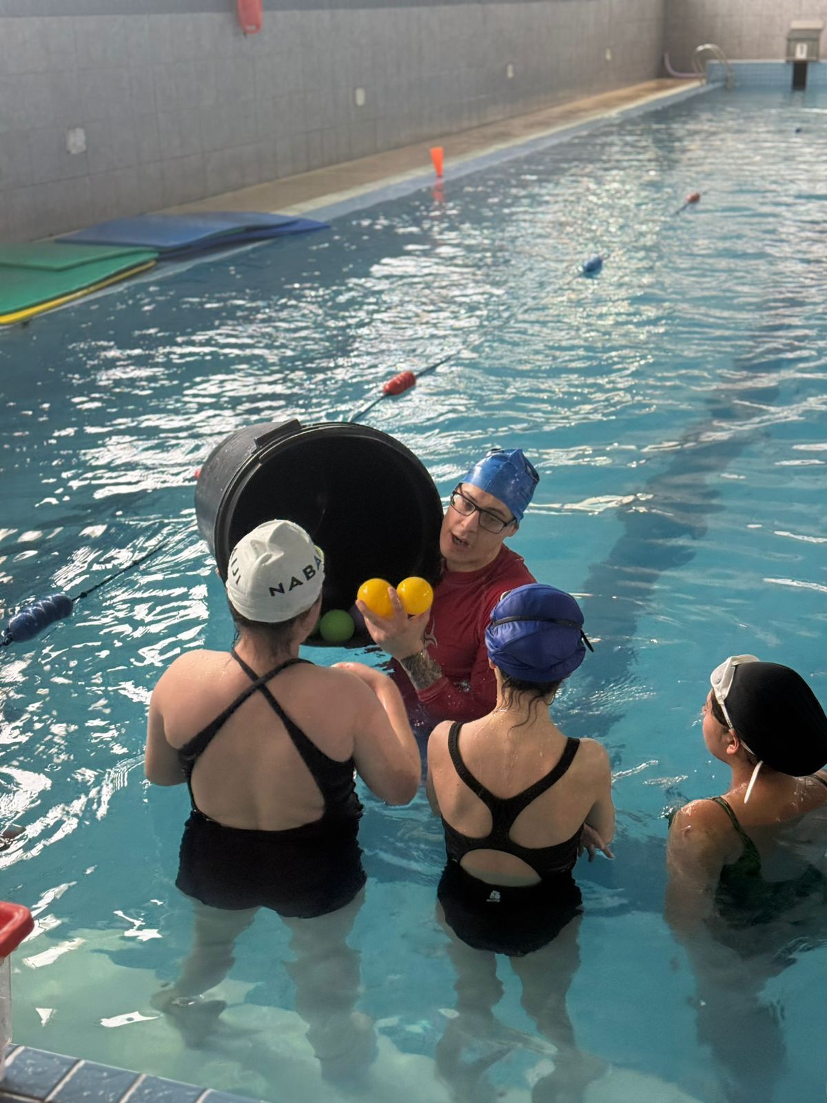 Personas en una piscina, una de ellas sosteniendo un balde y pelotas amarillas, posiblemente enseñando una clase de ejercicios acuáticos.