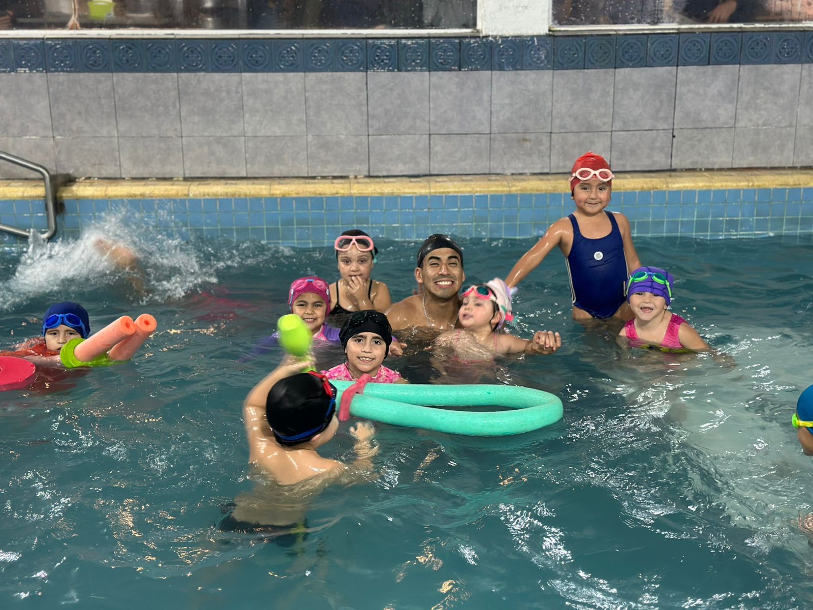 Niños y un instructor jugando en una piscina. Algunos niños usan gorros de baño. La piscina tiene azulejos azules y agua.