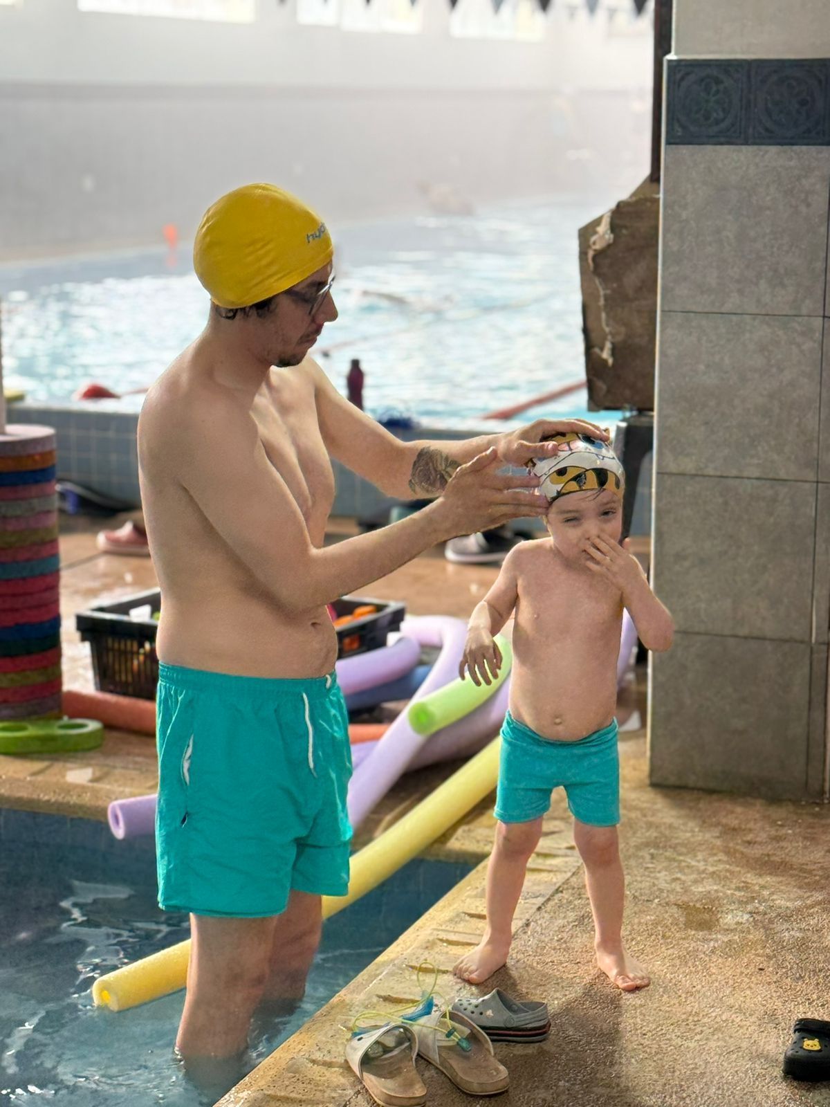 Un hombre con pantalones cortos color verde azulado y gorra amarilla ayuda a un niño con un gorro de baño en una piscina.