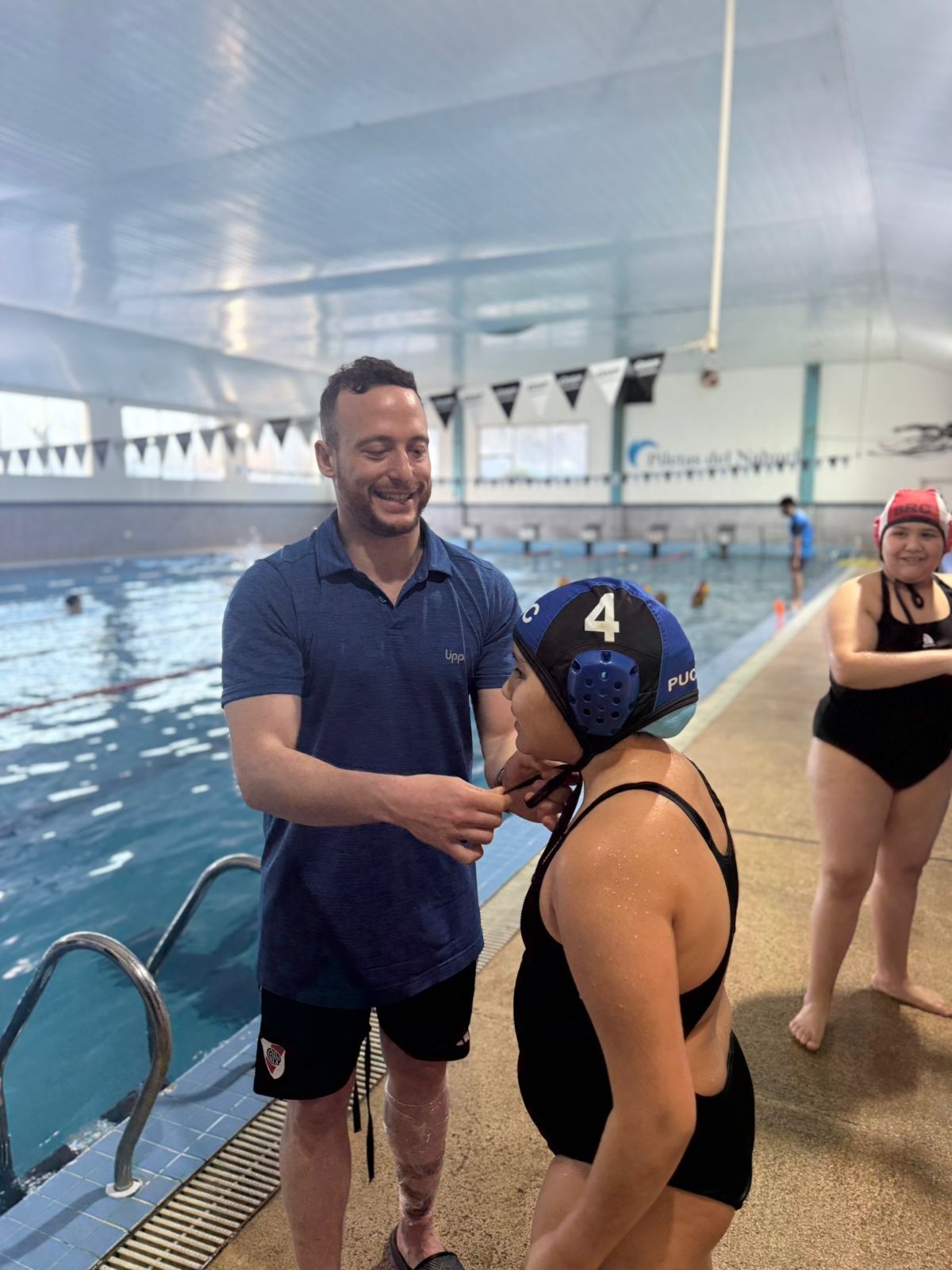 Hombre ajustando la gorra de waterpolo de una persona en traje de baño junto a la piscina.