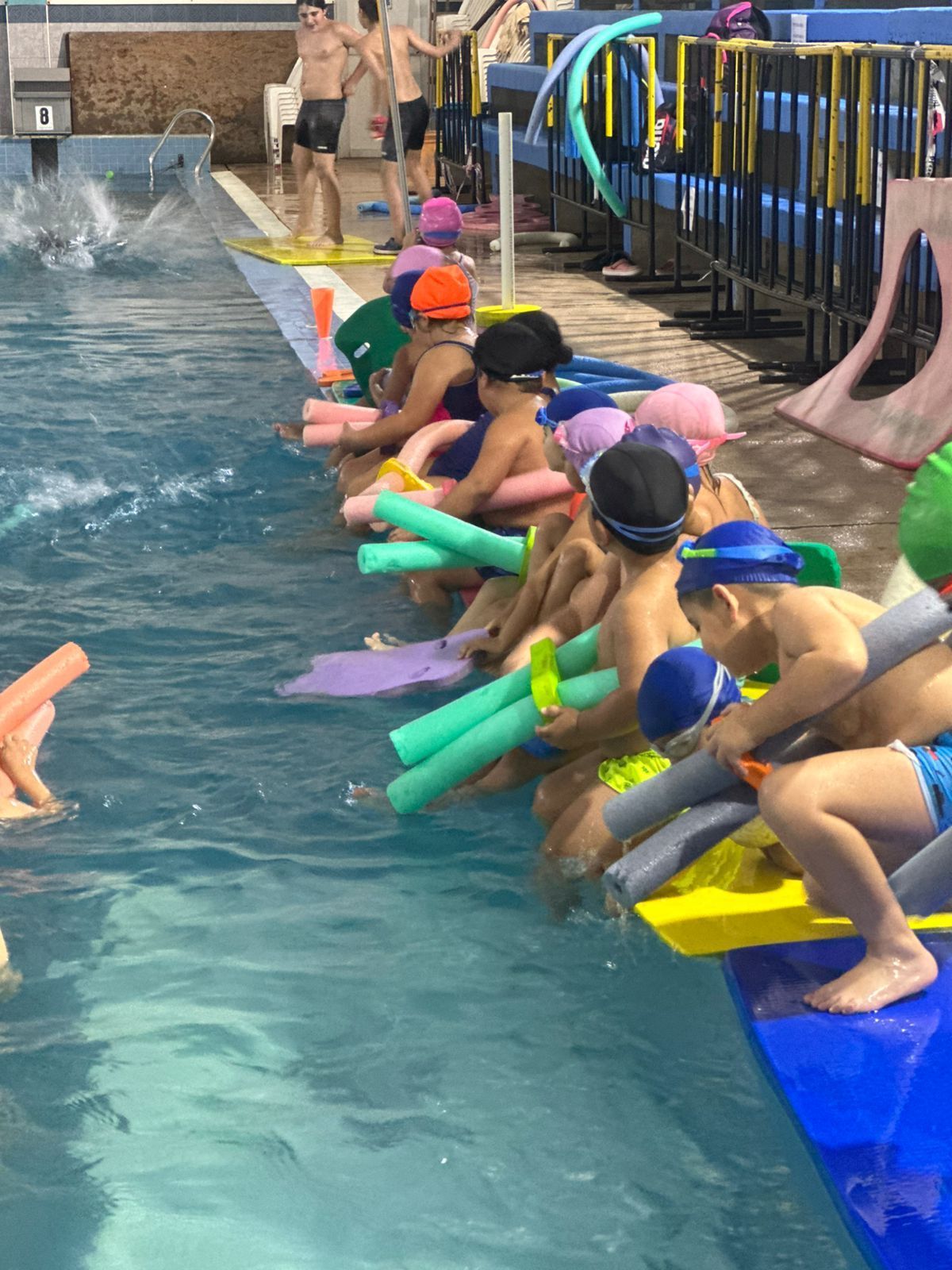 Niños en traje de baño se sientan en el borde de una piscina con flotadores. Hay otras personas en el agua.