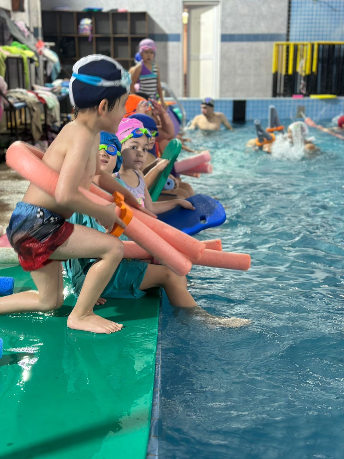 Niños en traje de baño en el borde de la piscina con fideos de piscina; otras personas nadan en el fondo.