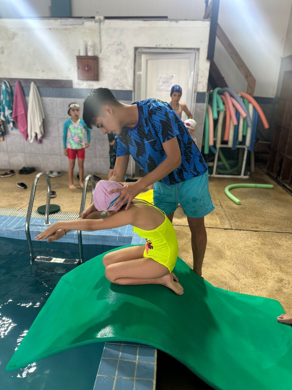 Un instructor de natación ayuda a un niño en una piscina. El niño se arrodilla sobre una plataforma verde, con un traje de baño amarillo y una gorra rosa.