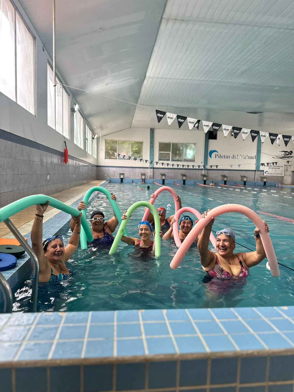 Personas en una piscina sosteniendo fideos de espuma de colores, haciendo ejercicio en un centro acuático interior.