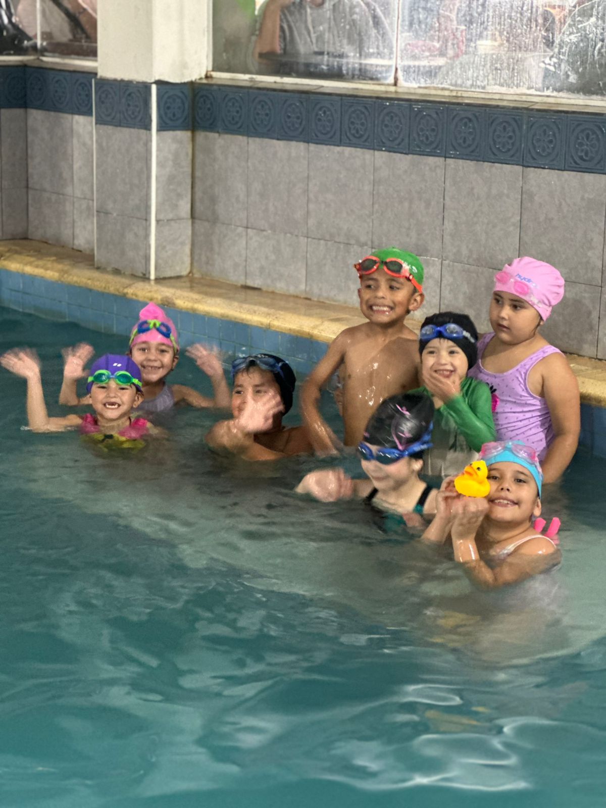 Grupo de niños en una piscina, sonriendo y saludando. Algunos llevan gorro y gafas de natación.