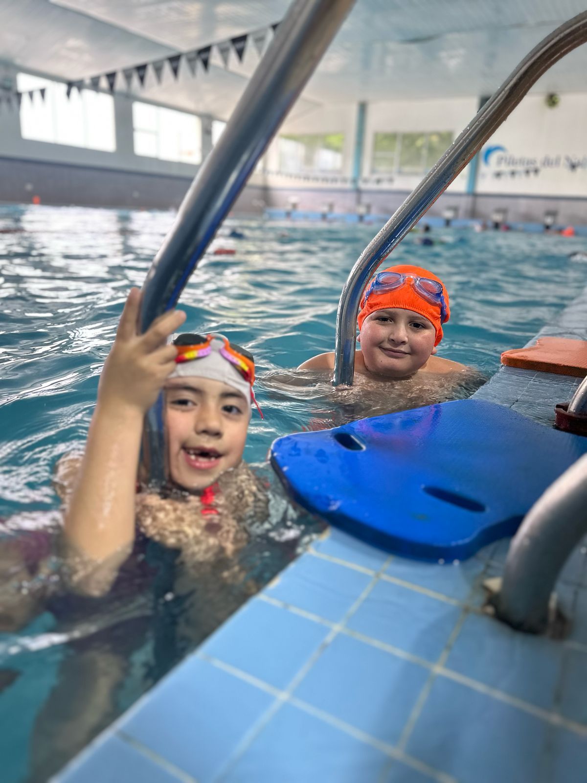 Dos niños con gorros y gafas de baño sonríen desde una piscina, agarrándose a una escalera.