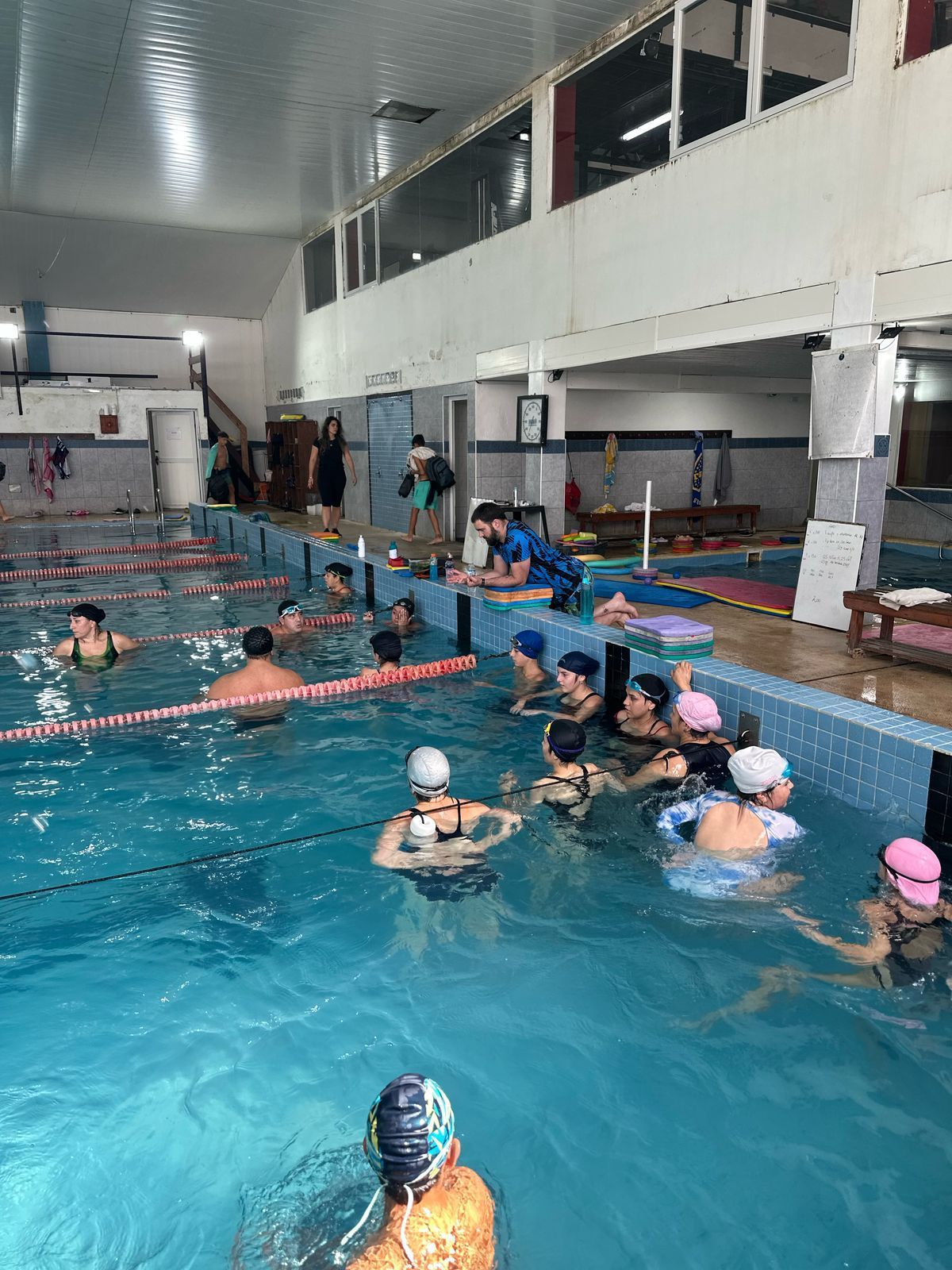 Personas en una piscina azul, nadando y haciendo ejercicio. Interior con paredes blancas, ventanas y algunos aparatos.