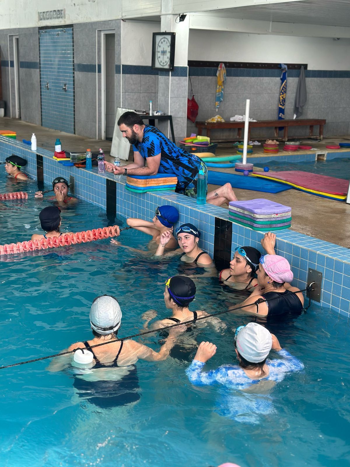Personas en una piscina, bajo la supervisión de un entrenador. Muchos llevan gorro de baño; ambiente interior.