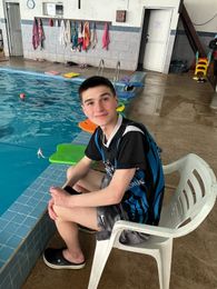 Un joven sonríe, sentado en una silla blanca junto a una piscina.