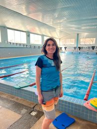 Mujer de pie junto a una piscina, sonriendo. Lleva camisa azul y pantalones cortos naranjas/grises. Piscina cubierta con carriles marcados.