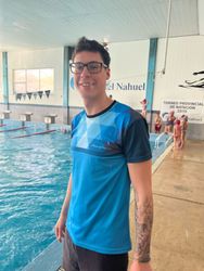 Un hombre con gafas y camisa azul sonríe junto a una piscina. Tatuaje en el brazo. Niños al fondo.