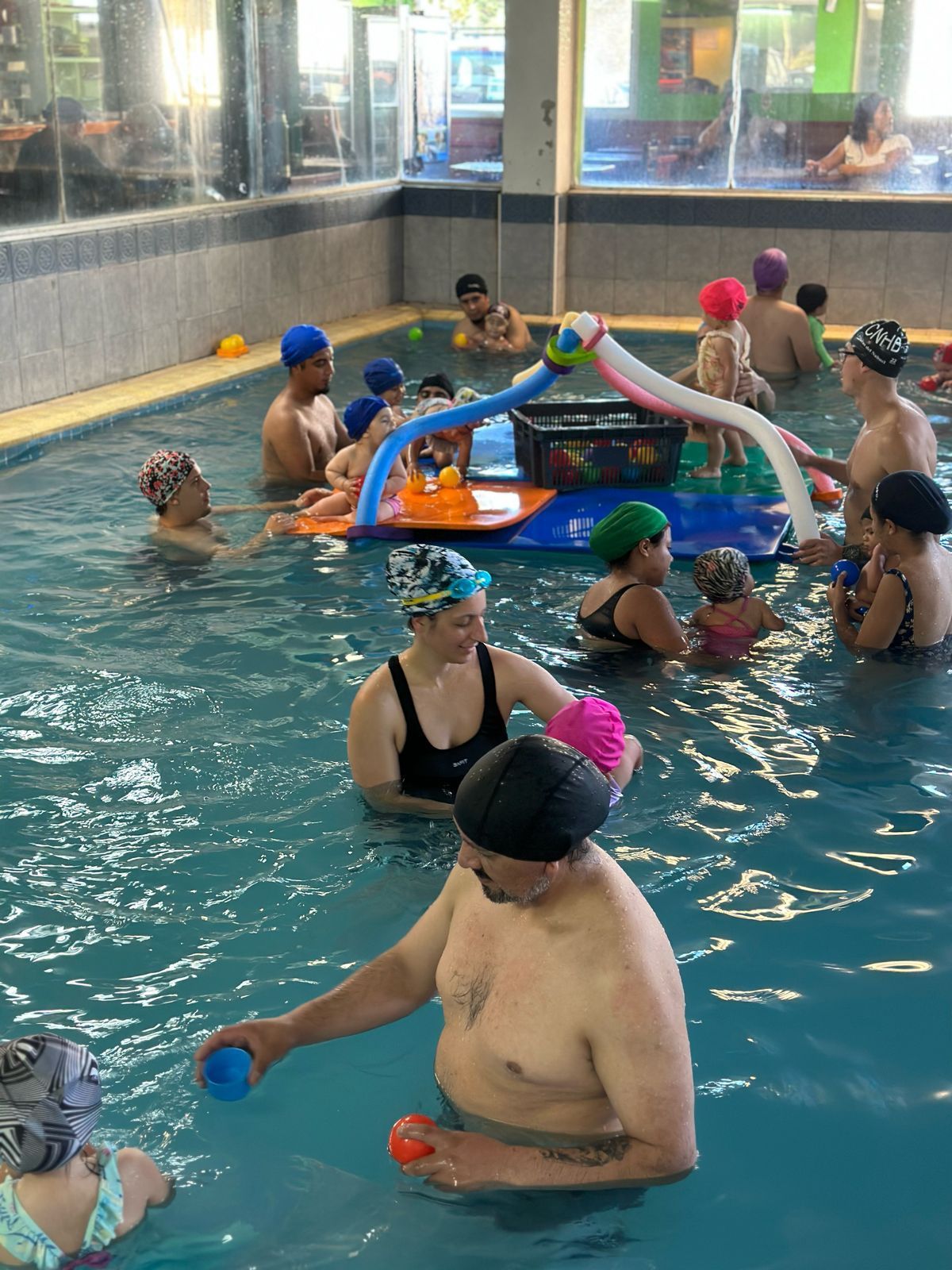Personas en una piscina con gorros de baño. Varios niños juegan con adultos cerca de un juguete acuático flotante.