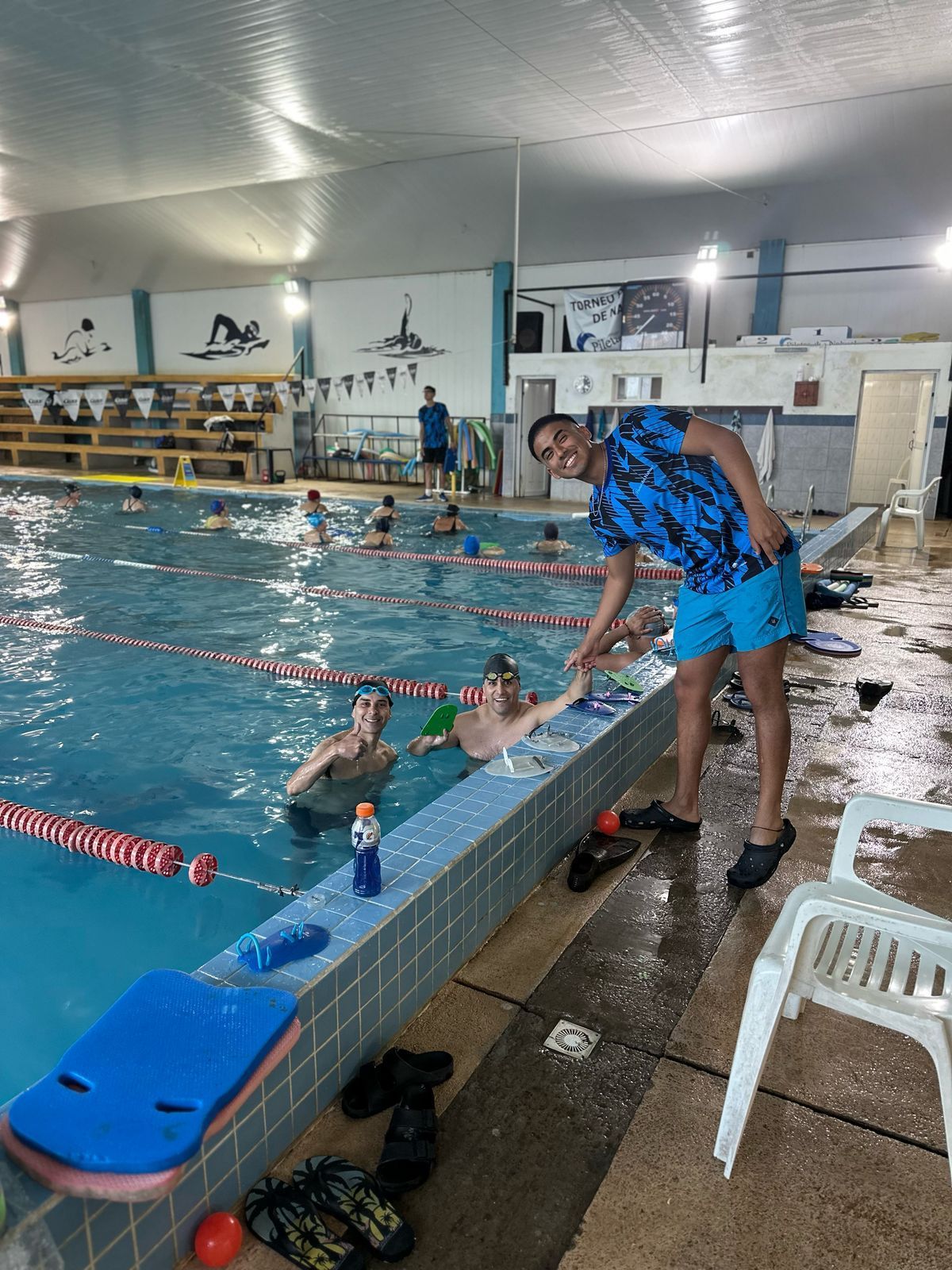 Personas en la piscina con el instructor. Este le extiende la mano a un nadador. Silla blanca y ayudas de natación azules y rojas.