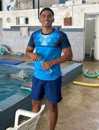 Hombre sonriendo, con una botella de agua en la mano, junto a la piscina. Viste camisa azul y pantalones cortos en una piscina cubierta.
