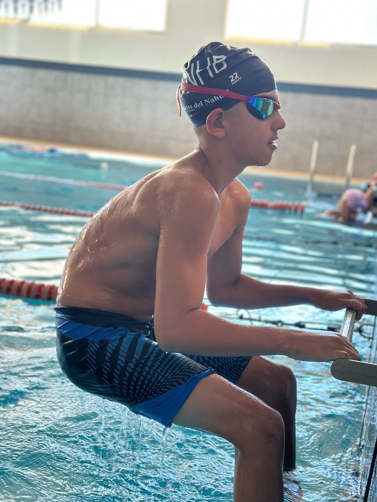 Nadador aferrado a una escalera de piscina, con gorro y gafas de natación. Pantalón corto azul y negro. Escenario de piscina.