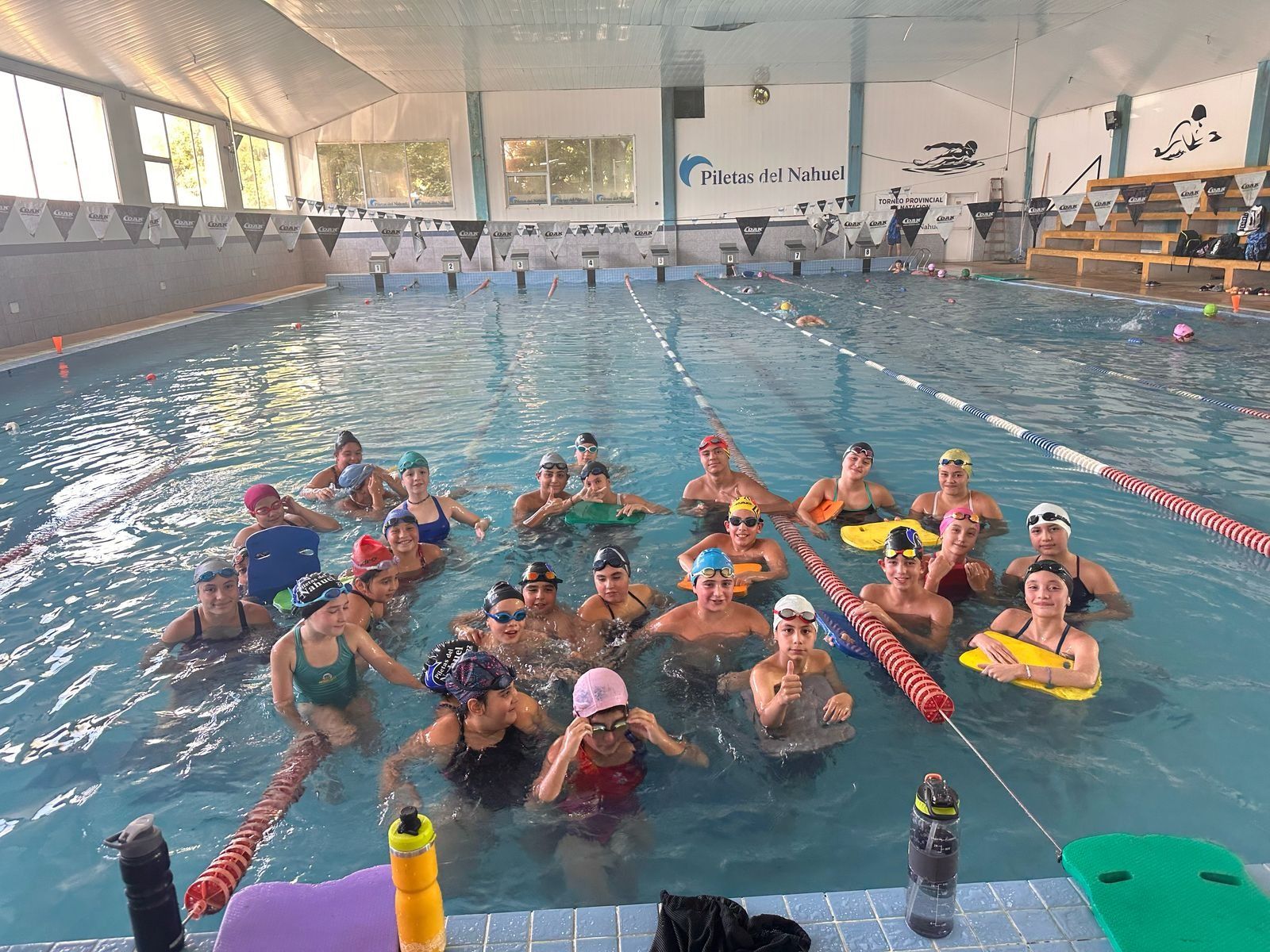 Grupo de personas en una piscina, algunas con dispositivos de flotación, bajo un techo blanco.