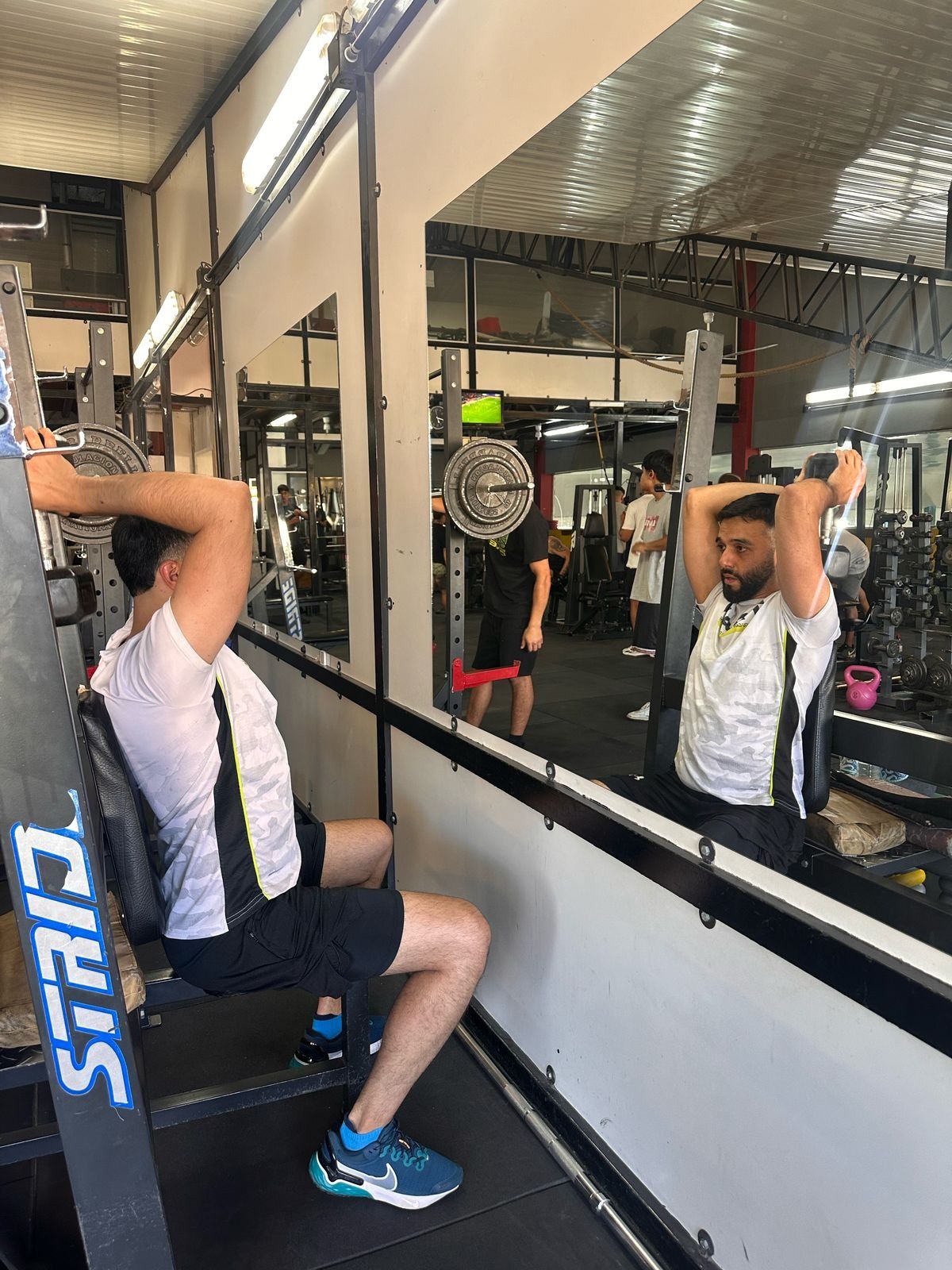 Hombre ejercitando tríceps con mancuernas en un gimnasio, visto desde un espejo. Lleva camisa blanca y pantalones cortos negros. Otras personas al fondo.