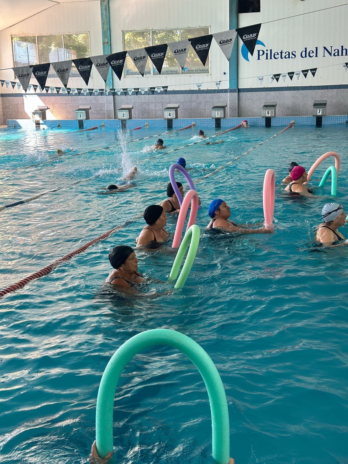 La gente en una piscina usa flotadores. La piscina es de agua azul y está cubierta, con una pancarta.