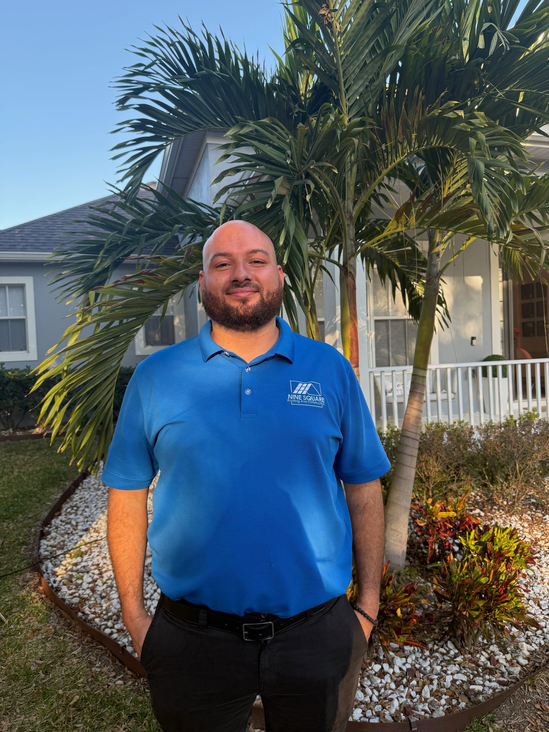 Man in blue polo shirt standing outside a house with a palm tree, smiling.