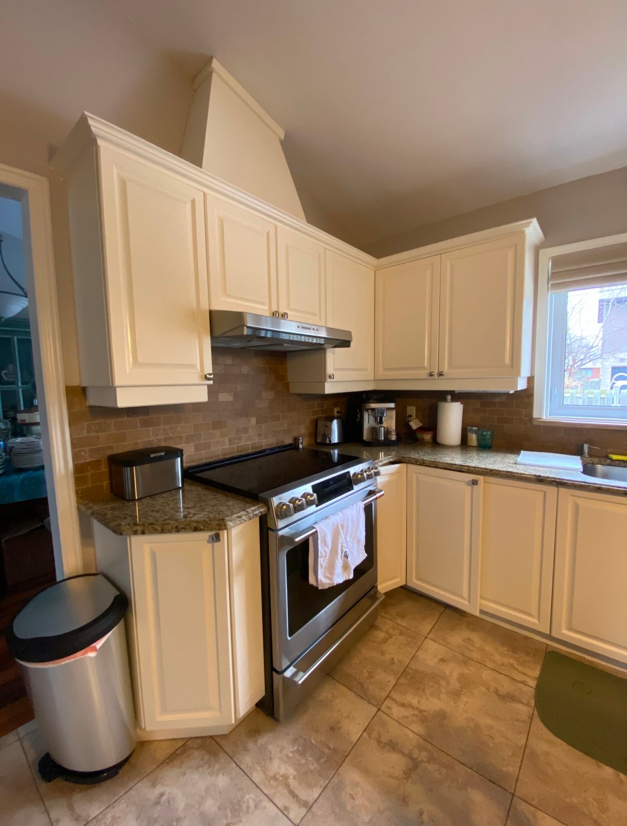 A kitchen with white cabinets , a stove , a sink and a trash can.