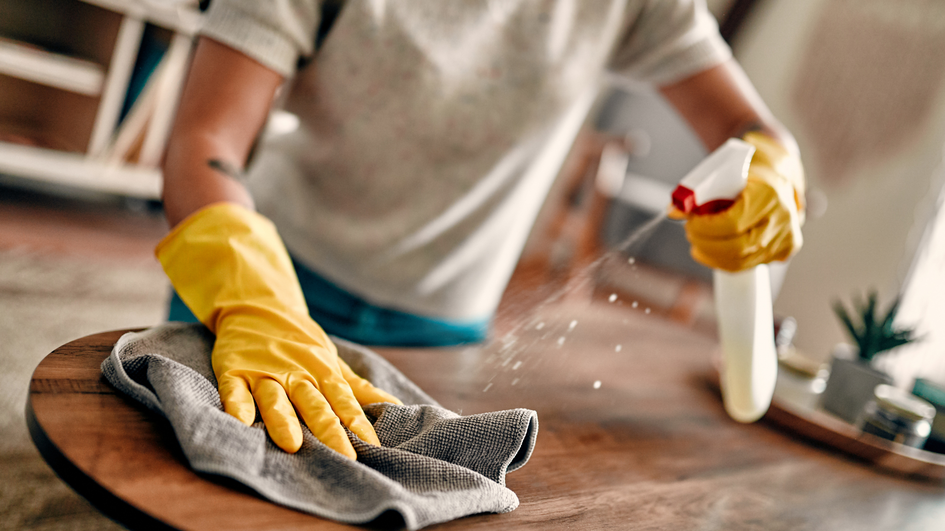 Une personne portant des gants jaunes nettoie une table en bois avec un chiffon et un vaporisateur.