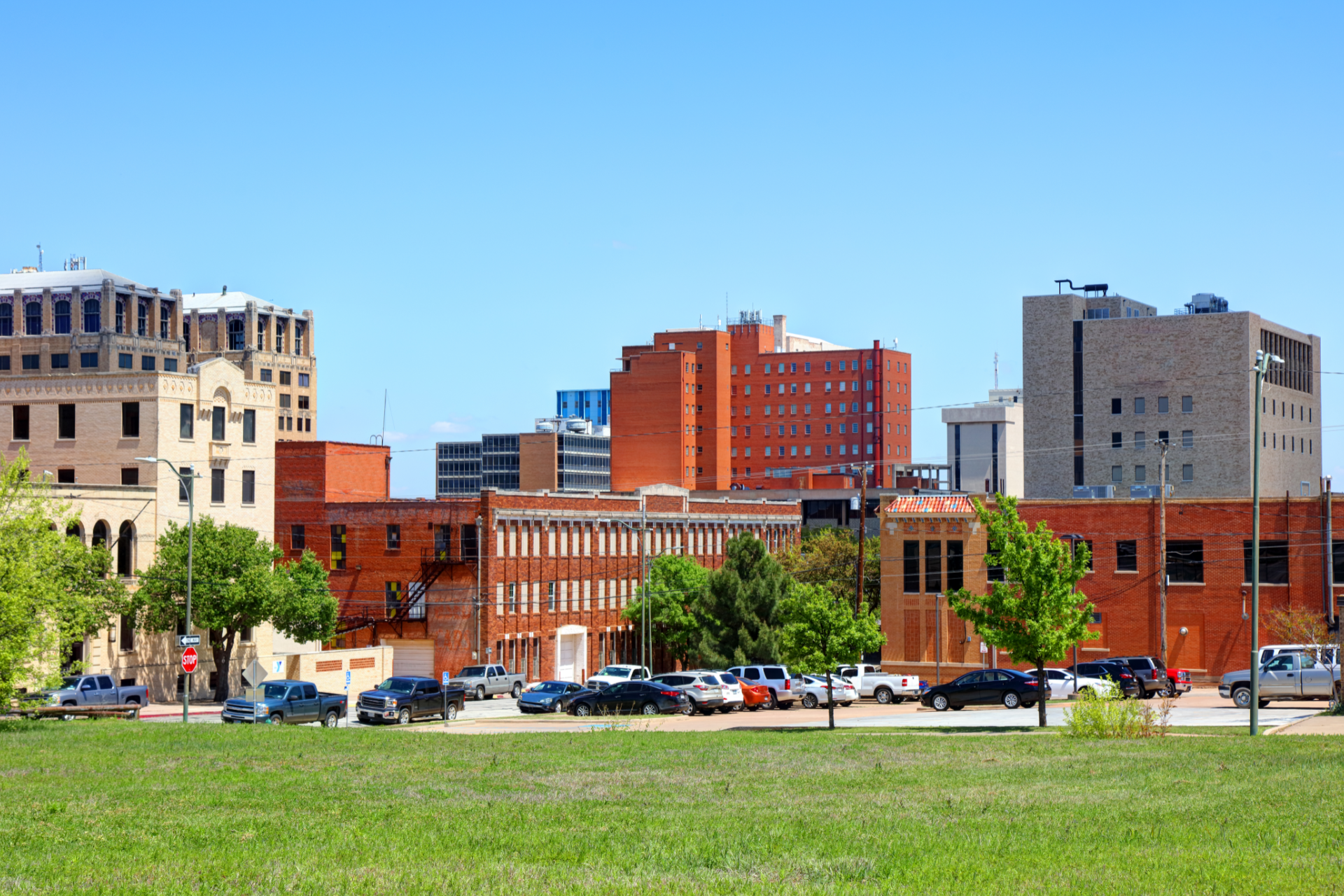 City skyline with brick buildings, parked cars, and a grassy foreground under a clear blue sky
