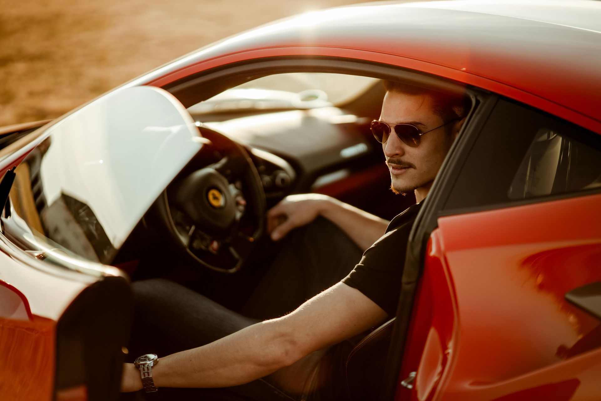 Man in sunglasses sitting in a red sports car with the door open on a sunny road