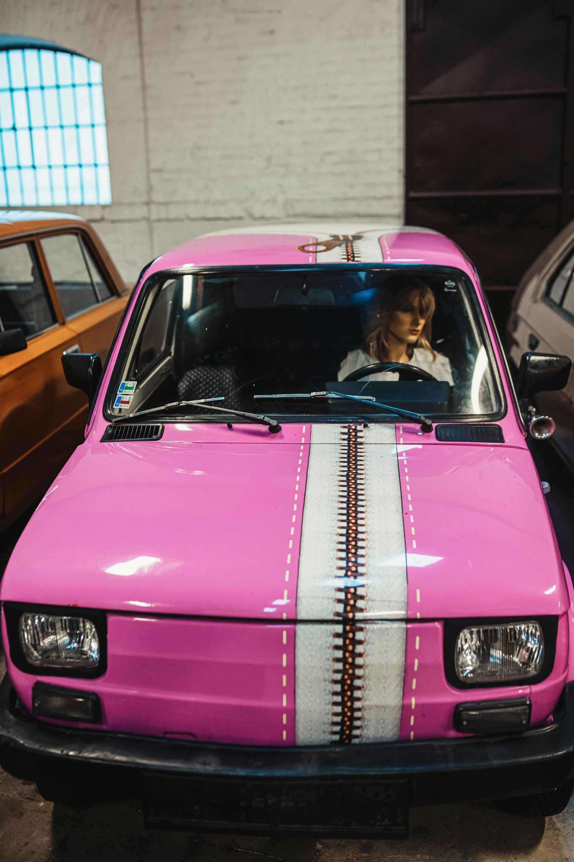 Pink car with a white center stripe parked indoors, driver visible through the windshield.