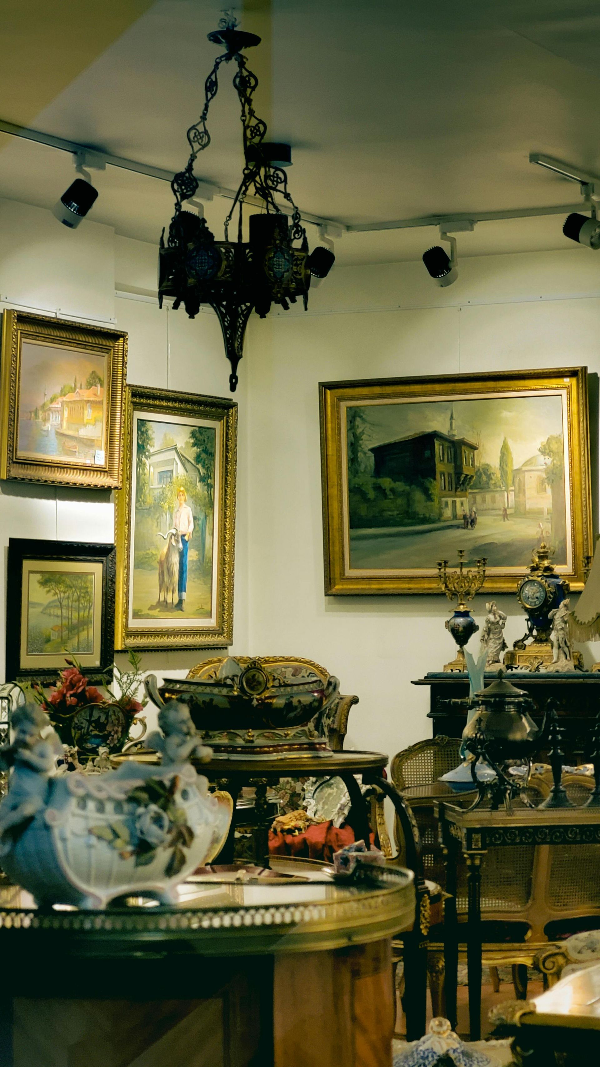 Antique-filled room with framed paintings, chandeliers, ornate furniture, and a decorative ceramic vase