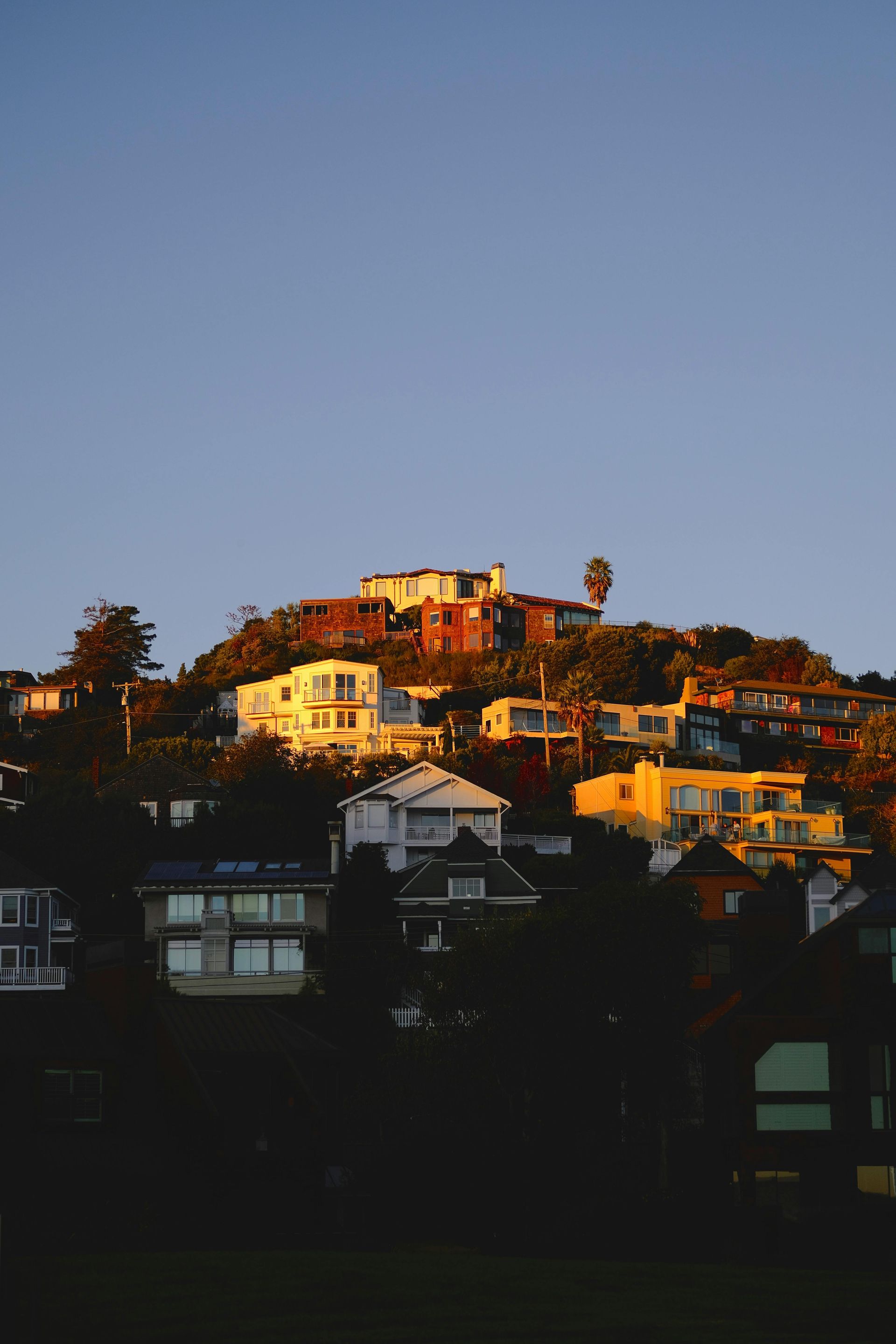 Sunlit houses on a hillside above dark rooftops at dusk under a clear blue sky